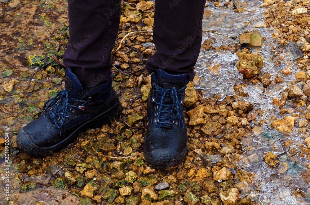 A lonely man in dark jeans and heavy road boots on lacing strollsstand  in the cold season on the river bank. On the shore, small stones, ice, thickets of reeds and shrubs.