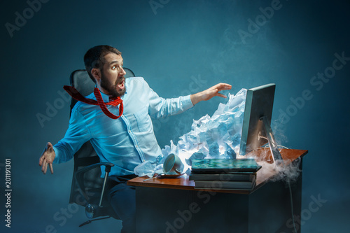 Young stressed handsome businessman working at desk in modern office shouting at laptop screen and being angry about spam