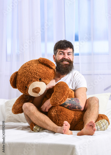 Guy on cheerful face hugs giant teddy bear. Birthday concept. Macho with beard and mustache cuddling with plush toy on birthday morning. Man sits on bed and hugs big toy white curtains
