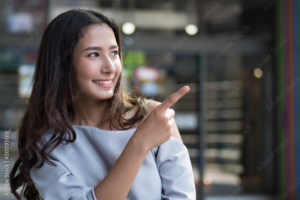 happy girl pointing up; portrait of cheerful smiling woman points hand and finger up to something; positive relaxed happy asian woman smiles in outdoor city scene; asian woman young adult model