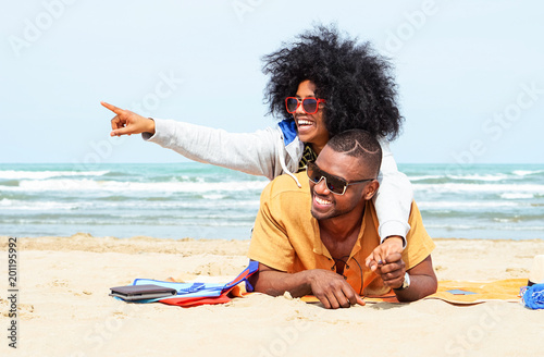 Young afro american couple  pointing finger relaxing on beach - Cheerful african friends having fun at day on blue ocean background - Concept of lovers happy moments on summer holiday