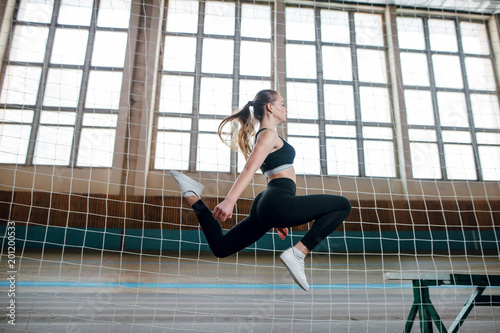 Portrait of a young fit gymnast woman in black sportswear working out in the old stadium. Beautiful blonde girl jumping and doing gymnastic poses. Window background. Indoors