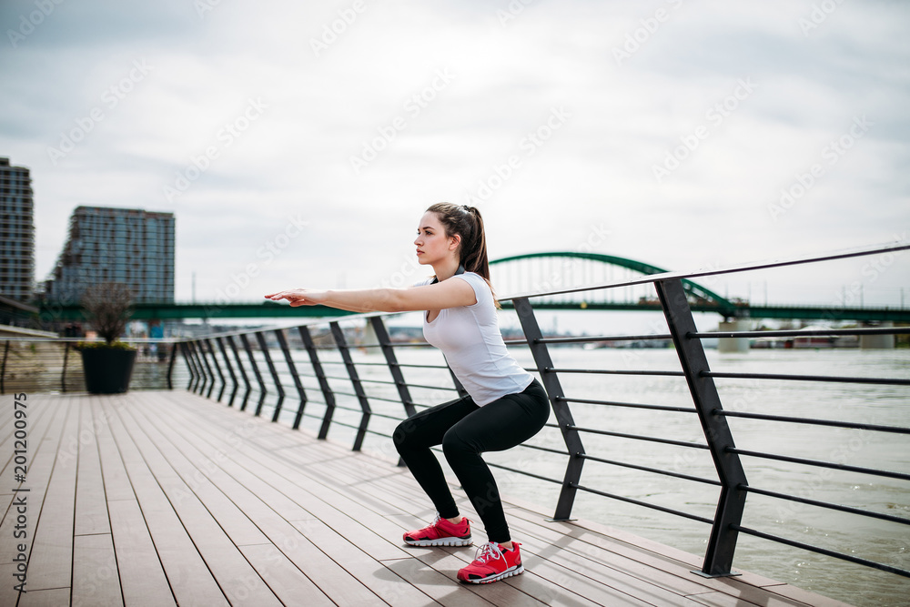 Side portrait of young woman doing squat exercises on a river bank.