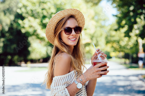 Smiling attractive girl in sunglasses holds a fresh cocktail, looking at camera, wearing trendy clothes and hat. Summertime.