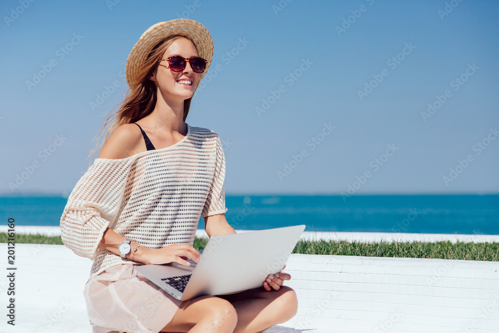 Attractive cheerful girl in sunglasses using a laptop, while sitting on quay, near the sea. Dressed in fashionable clothes, in summer hat. Outdoors.