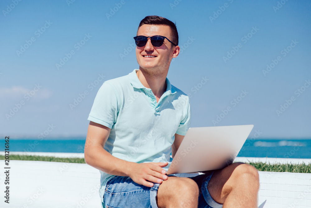 Smiling attractive man in sunglasses with laptop, looking away, while sitting on quay, near the ocean, wearing t-shirt and jeans shorts.