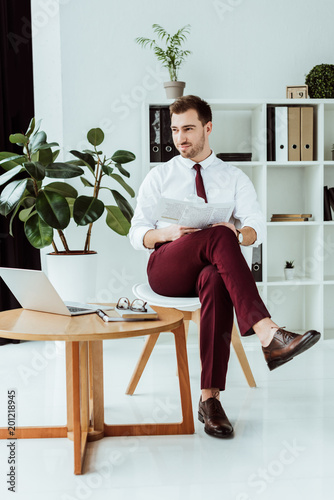 handsome stylish businessman with newspaper and laptop sitting in modern office