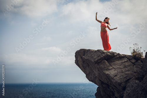 A young woman with beautiful forms and thin waist dancing dance of freedom on a cliff above a blue, deep sea. Femininity. Wildness. Naturalness. Wind. Wild life. Incredible landscape. Happiness.