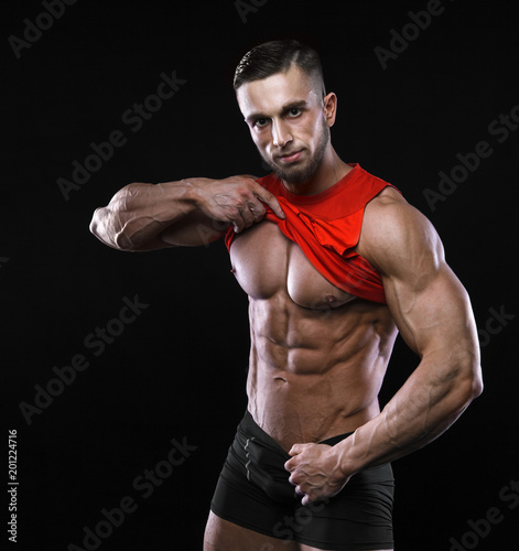 Cute young sports man in red t-shirt shows relief abdominal muscles in gym