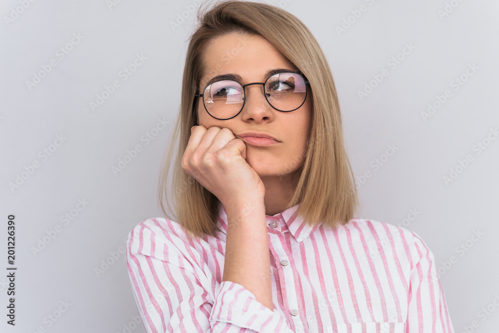Studio portrait of sad blonde female student wearing pink shirt and round spectacles, sitting with unhappy boring expression on her face, resting chin on hand listening information. People, emotion