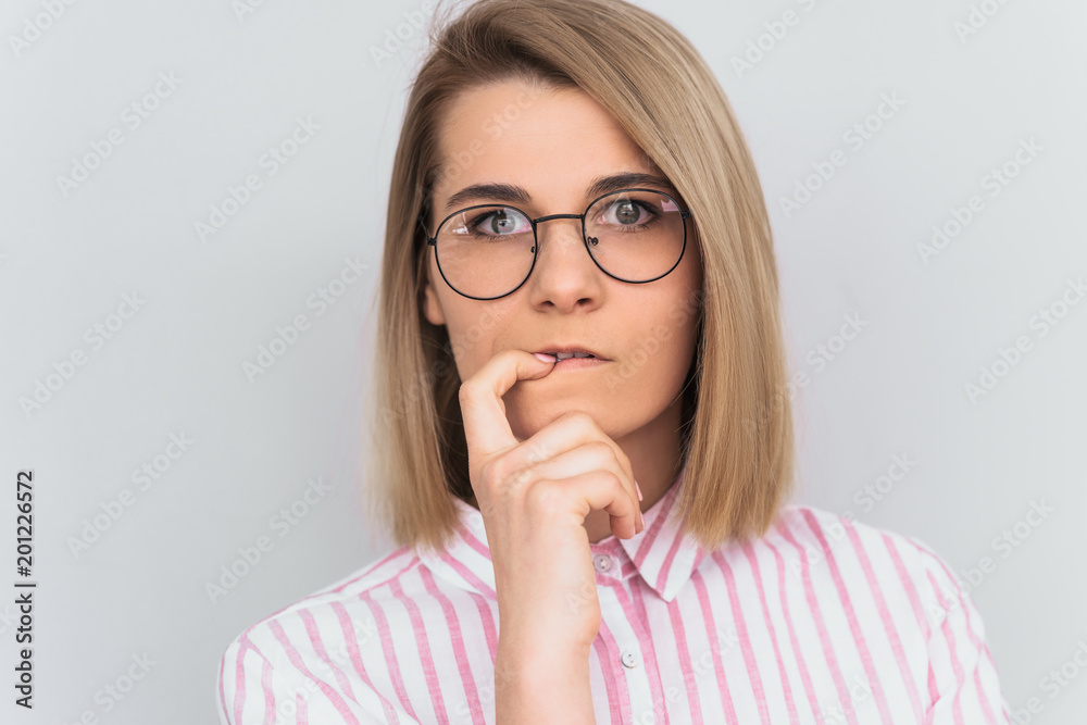 Closeup shot of serious attractive young European woman wearing pink shirt and round glasses keeping finger on her lips, having thoughtful indecisive look, thinking about something important. People