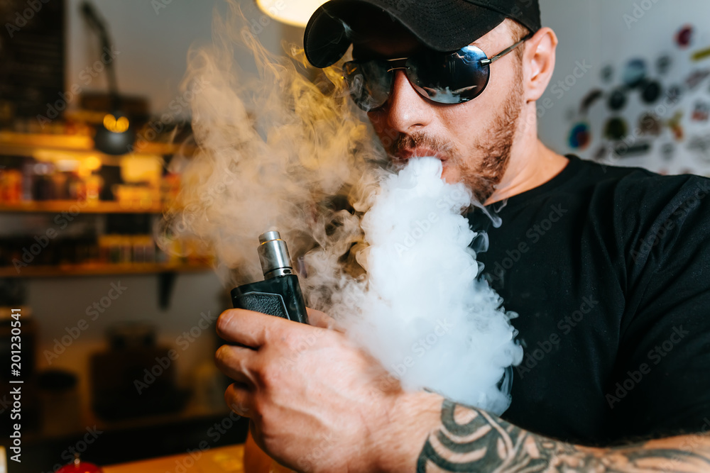 Brutal bearded man in sunglasses smoke an electronic cigarette and releases clouds of vapor at the vape shop. Brutal bearded man in sunglasses smoke an electronic cigarette and releases clouds of vapor at the vape shop.