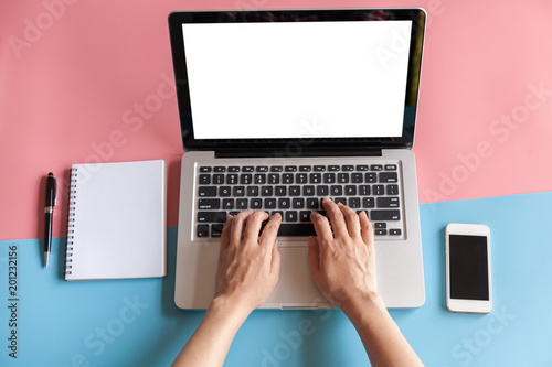 Top view of woman’s hands using laptop with notebook, pen and smartphone on pink and blue pastel color background.
