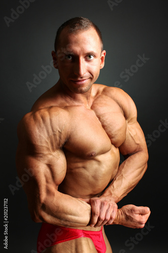 Studio shot of young male bodybuilder posing, showing his front chest muscles and huge biceps, on black background.
