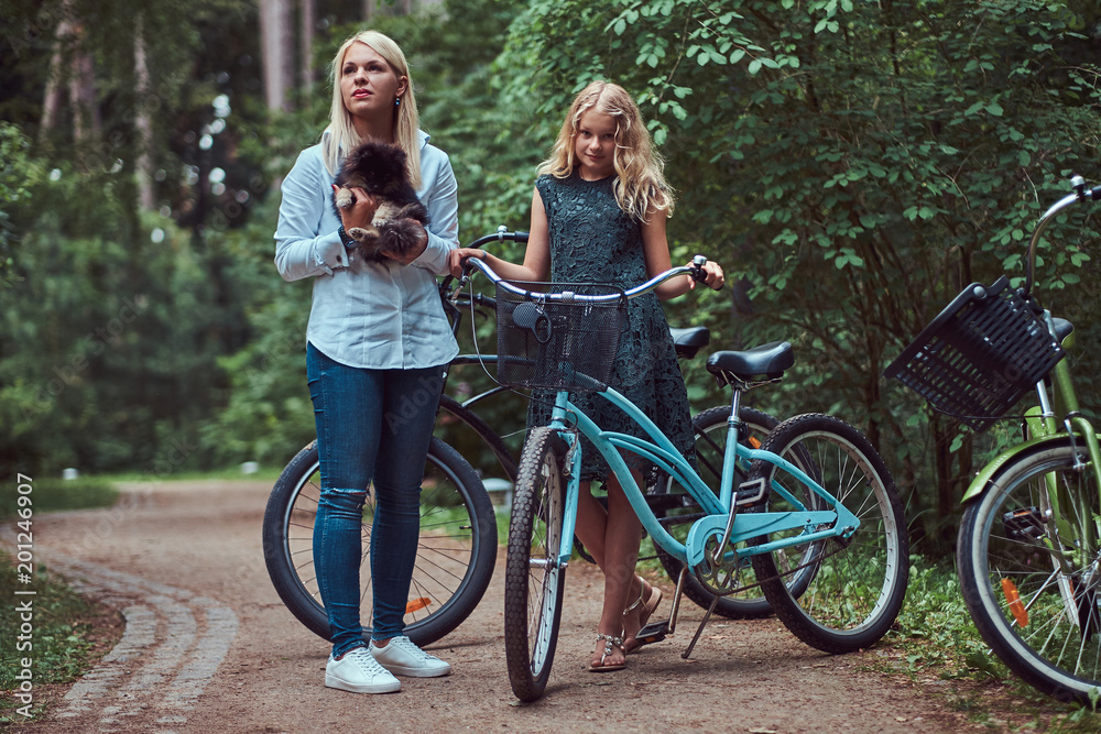 Portrait of a mother and daughter with a blonde hair on a bicycle ride with their cute little spitz dog in the park. Portrait of a mother and daughter with a blonde hair on a bicycle ride with their cute little spitz dog in the park.