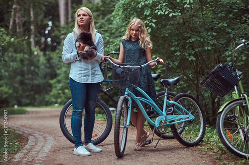 Portrait of a mother and daughter with a blonde hair on a bicycle ride with their cute little spitz dog in the park.