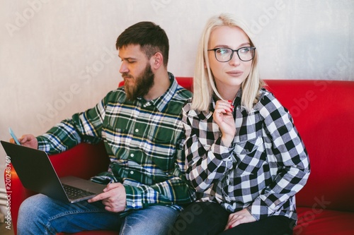 Blonde girl with glasses sits with a guy in a green shirt on a red couch with a laptop