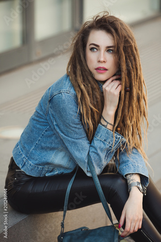 Stylish woman with dreadlocks sitting on street in denim jacket