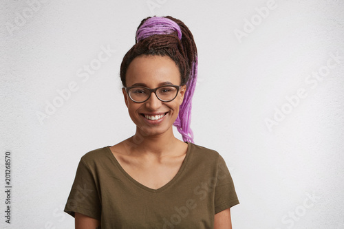 Indoor shot of young glad mixed race woman, weras casual t-shirt and spectacles, dyed dreadlocks smiles and feel happy. Portrait of joyful african american photo