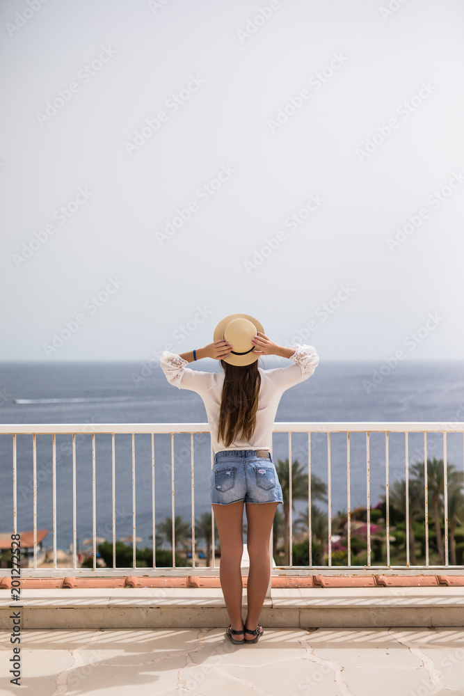 A beautiful young girl in a straw hat standing on a summer terrace and looking at the ocean at terrace