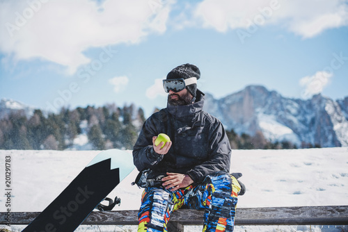 Bearded snowboarded in sunglass mask, at the ski resort on the background of mountains and blue sky.Man taking rest and holding green apple on hand. Horizontal.Blurred background