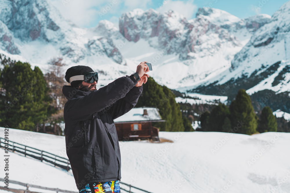 Bearded young snowboarded in sunglass mask, at the ski resort on the background of mountains and blue sky.Man making selfie photo on his smart phone. Horizontal.Blurred background