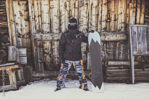Snowboarder wearing black jacket, mask with snowboard close authentic wooden house. Horizontal photo