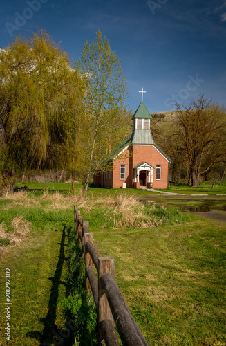 Wooden pole fence leads to a country church with willow tree and blue sky