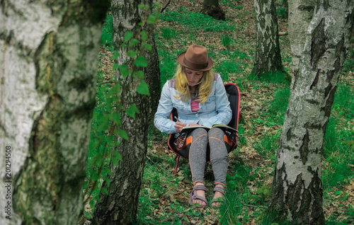 young caucasian female artist sitting outdoors in a forest and drawing a picture into her notebook