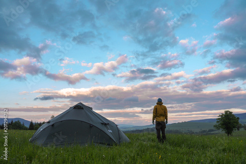 man standing next to a tent pitced on a meadow, looking at a clouds during sunset