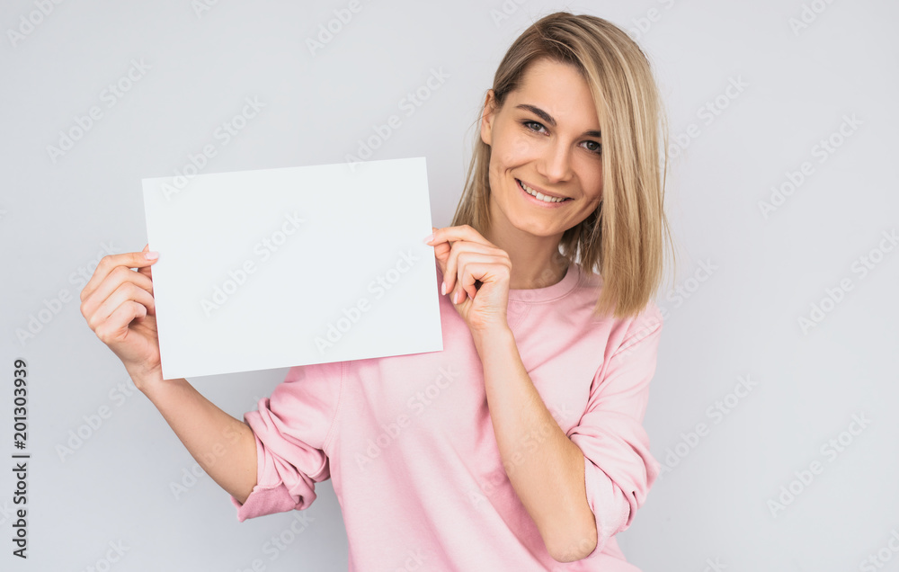 Closeup portrait of beautiful smiling blonde female wearing pink sweater, posing with white blank paper with copy space for your advertisement information and looking at camera, against white wall. Closeup portrait of beautiful smiling blonde female wearing pink sweater, posing with white blank paper with copy space for your advertisement information and looking at camera, against white wall.