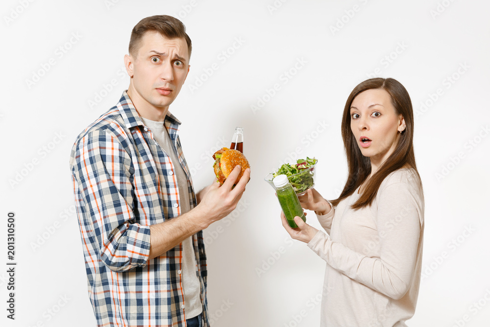 Couple man and woman standing with green detox smoothies, salad in glass bowl, burger, cola in glass bottle isolated on white background. Proper nutrition, healthy lifestyle, fast food, choice concept Couple man and woman standing with green detox smoothies, salad in glass bowl, burger, cola in glass bottle isolated on white background. Proper nutrition, healthy lifestyle, fast food, choice concept