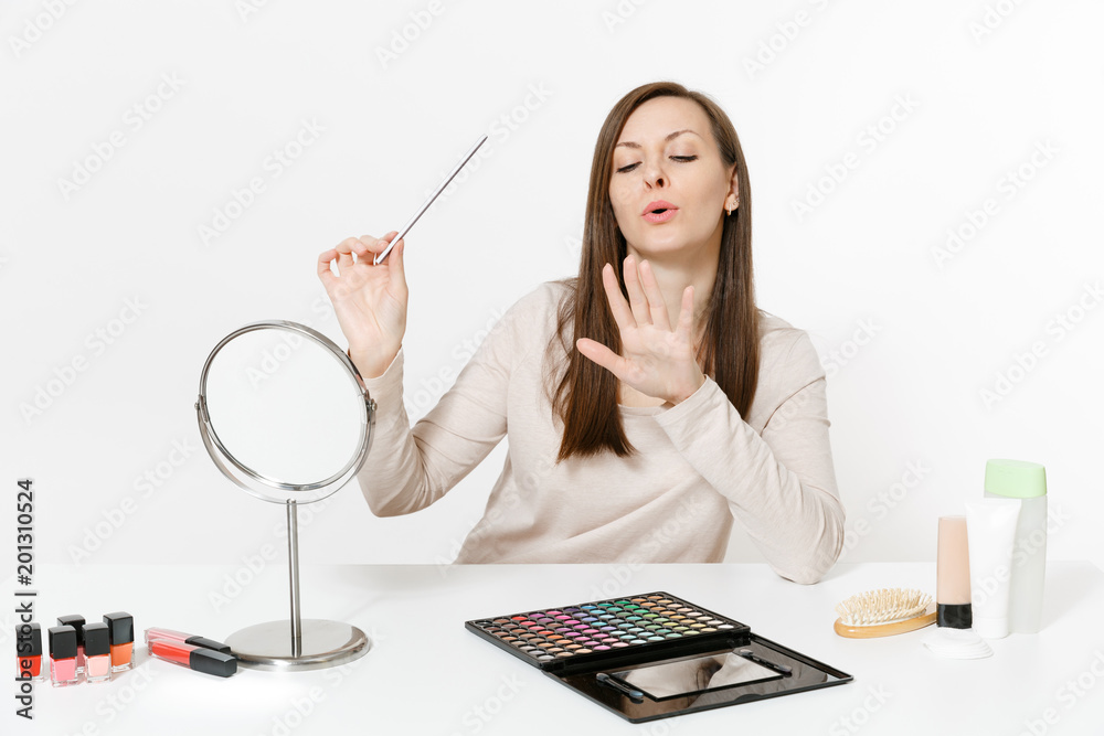 Attractive woman correcting fingernail using nail fail, sitting at table applying makeup with set facial decorative cosmetics isolated on white background. Beauty female fashion lifestyle concept.