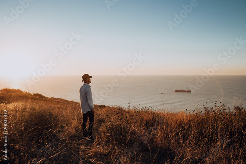 Man Standing on Coastal Cliff Overlooking Ocean at Sunset – Tranquil Outdoor Seascape with Golden Light and Distant Ship