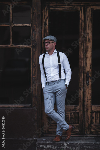 Fashionable retro dressed man with cap, suspenders and eyeglasses standing on city street and looking at side. Low angle view.