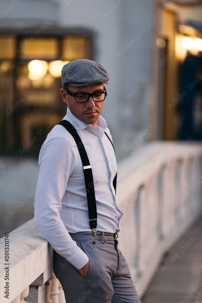 Fashionable retro dressed man with cap, suspenders and eyeglasses standing on city street and looking at side. Low angle view.