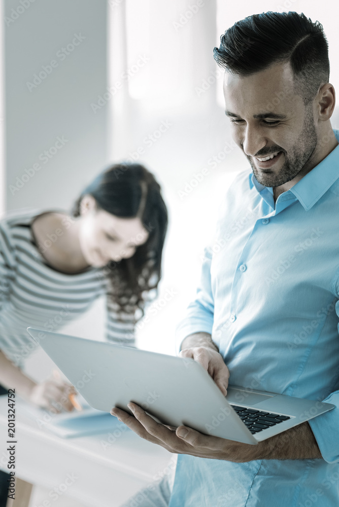 Business correspondence. Handsome appealing optimistic man carrying laptop while gazing down and grinning Business correspondence. Handsome appealing optimistic man carrying laptop while gazing down and grinning