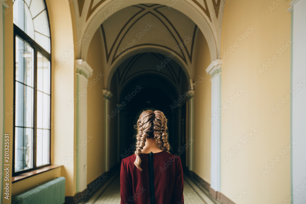 Back view of romantic blond woman student with cute hairstyle two braids wearing burgundy dress while standing indoor in hallway of old university building. Mood and study concept.