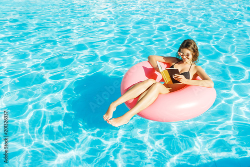 Canvas Print Beautiful happy woman reading a book with inflatable ring relaxing in blue swimm