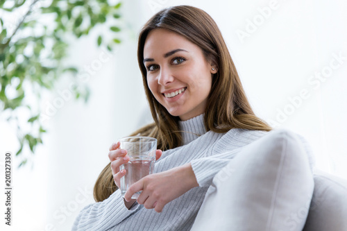 Beautiful young woman looking at camera while holding glass of water at home.
