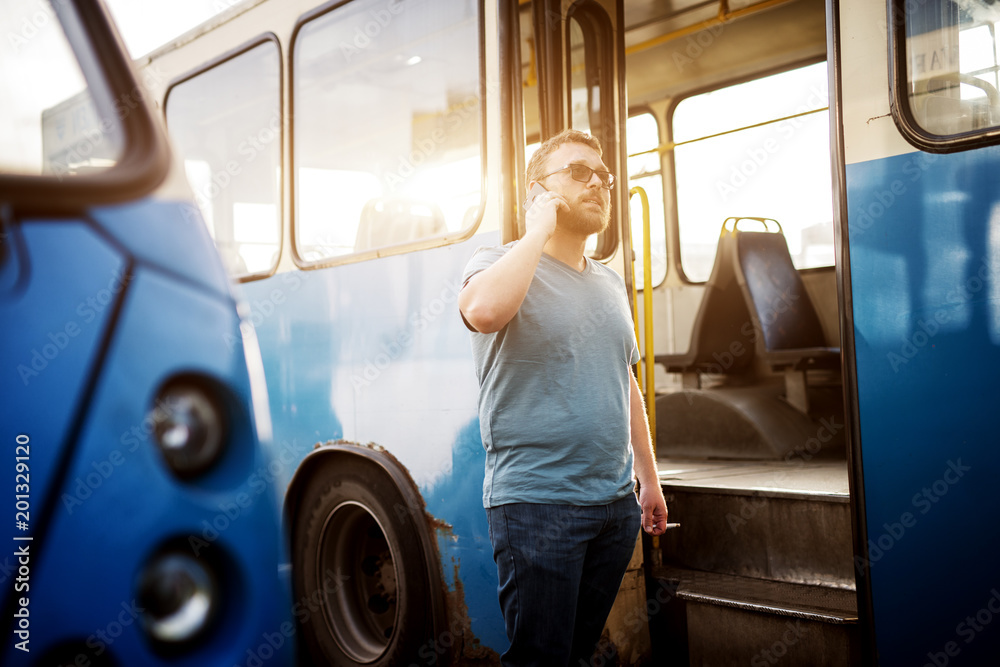 Young handsome man is talking on a phone and smoking a cigarette while standing outside the bus. Young handsome man is talking on a phone and smoking a cigarette while standing outside the bus.
