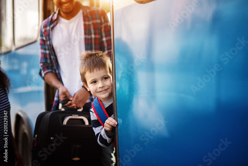 Little cheerful boy is peaking outside the blue bus while a young man is looking at him and smiling.