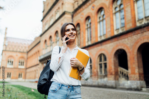 Foto Positive brunette woman in casual wear having mobile conversation at campus with book on sunny day