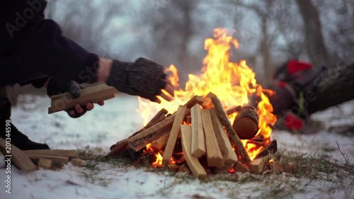 Portrait cropped of camper man in gloves and warm clothing putting wooden pieces into burning bonfire on snowy ground, slow motion
