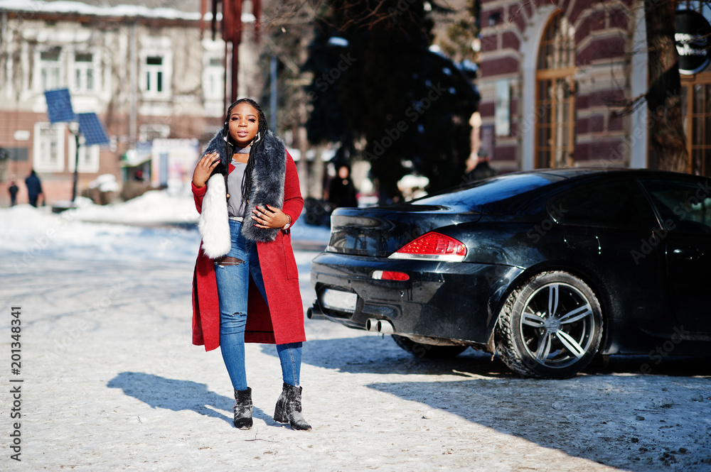 Rich african american girl in red coat and fur on winter street at sunny weather against black business car. Black stylish woman.