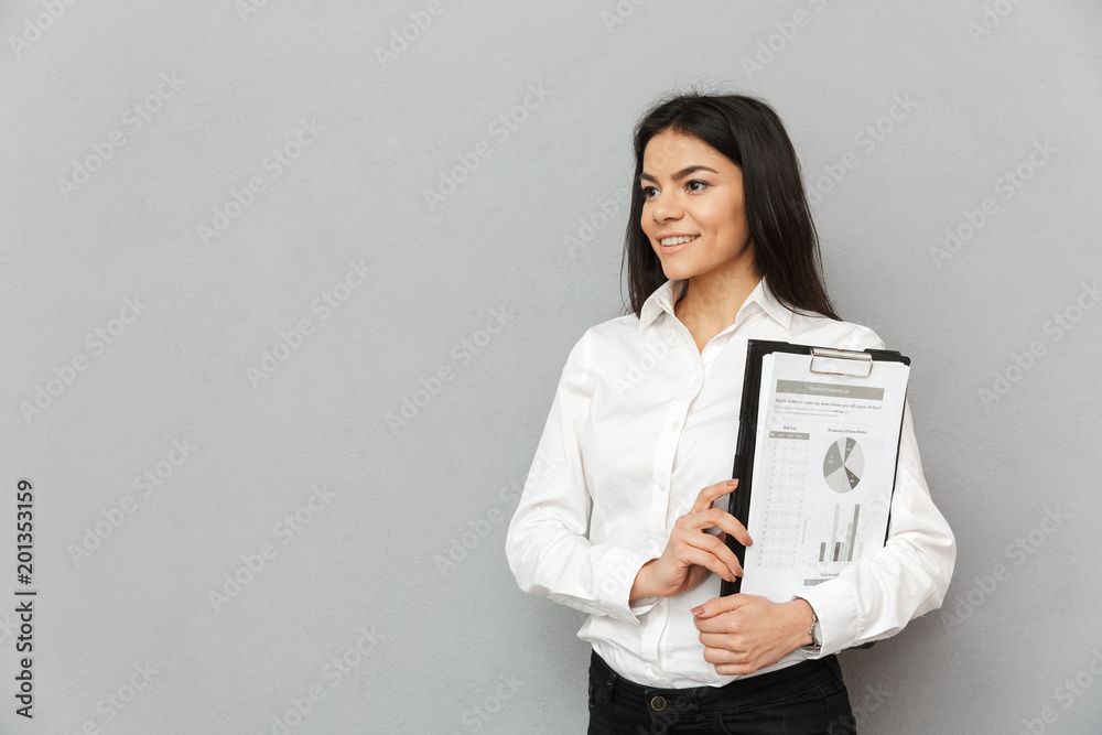 Photo of beautiful office woman wearing businesslike outfit holding clipboard with papers and looking aside, isolated over gray background Photo of beautiful office woman wearing businesslike outfit holding clipboard with papers and looking aside, isolated over gray background