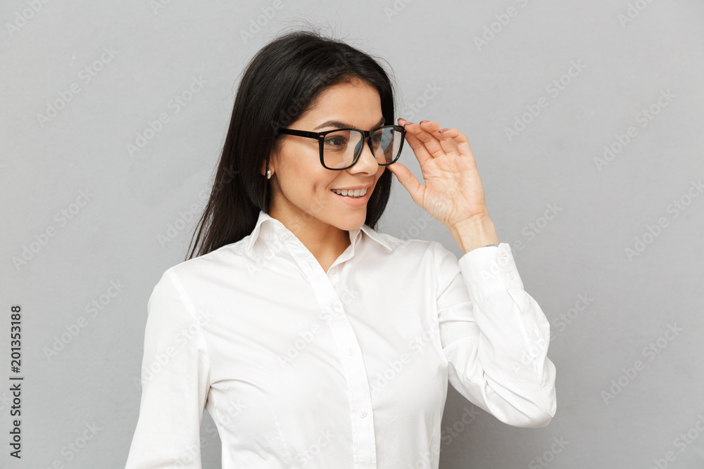 Portrait closeup of smiling successful woman with long brown hair wearing office clothing looking aside and touching her eyeglasses, isolated over gray background Portrait closeup of smiling successful woman with long brown hair wearing office clothing looking aside and touching her eyeglasses, isolated over gray background