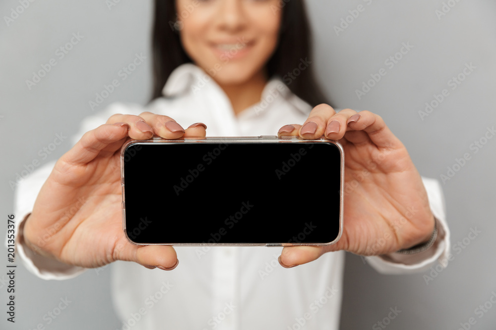 Photo cropped of brunette woman 30s in office wear holding cell phone in hands and demonstrating copyspace black screen on camera, isolated over gray background