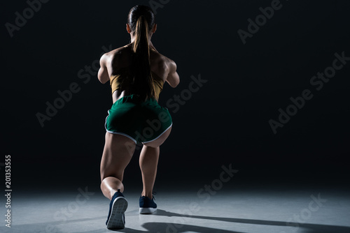back view of muscular sportswoman doing lunges, on black