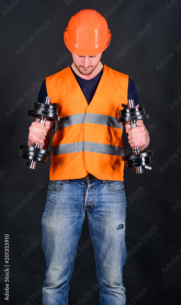Man in helmet, hard hat holds heavy dumbbells, black background. Worker, contractor, builder on busy face with muscular biceps. Strong builder concept. Builder in helmet working out, training.